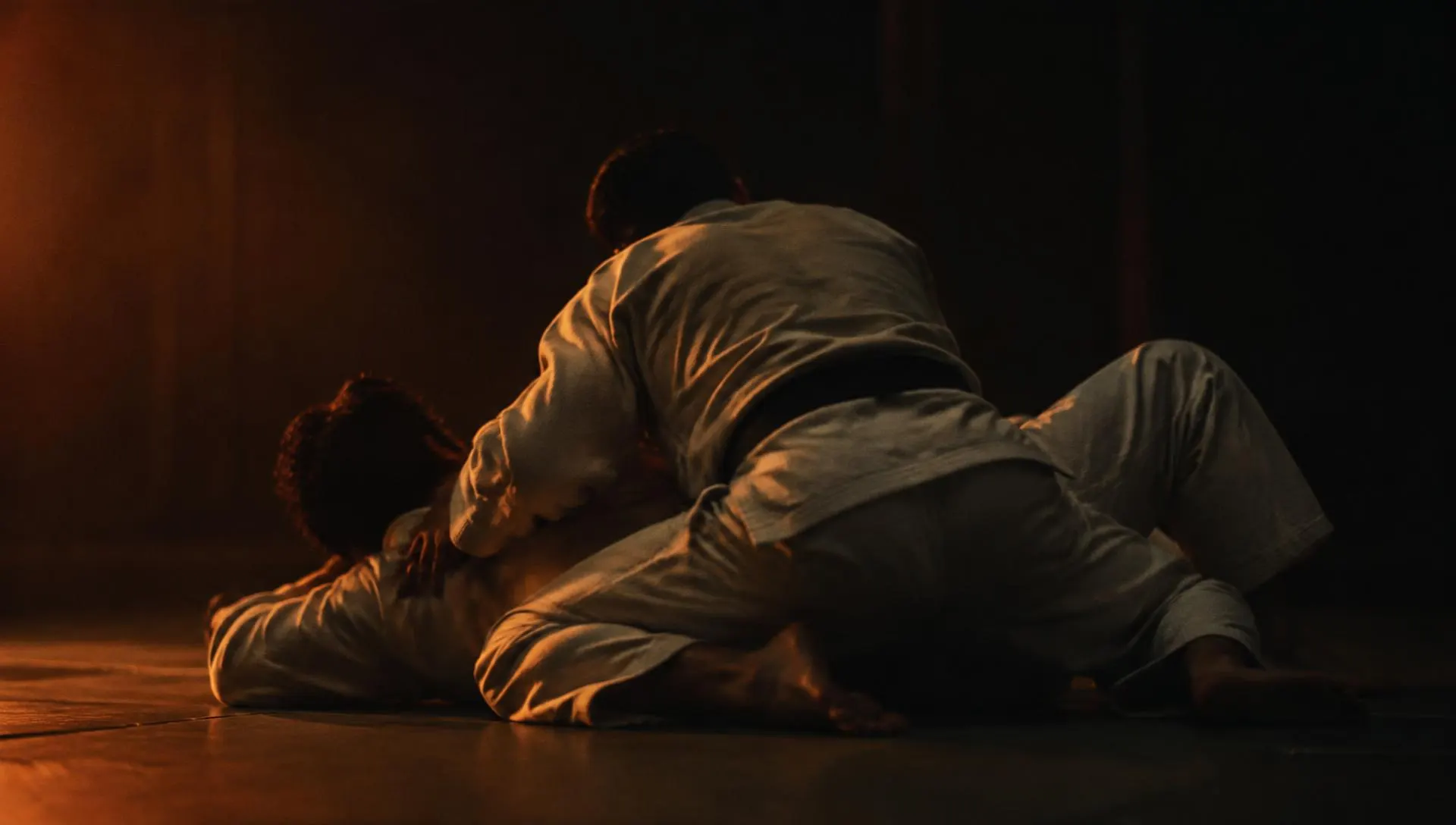Dois praticantes de Jiu-Jitsu de kimono a treinar num tatame em penumbra, iluminados por uma luz quente lateral.